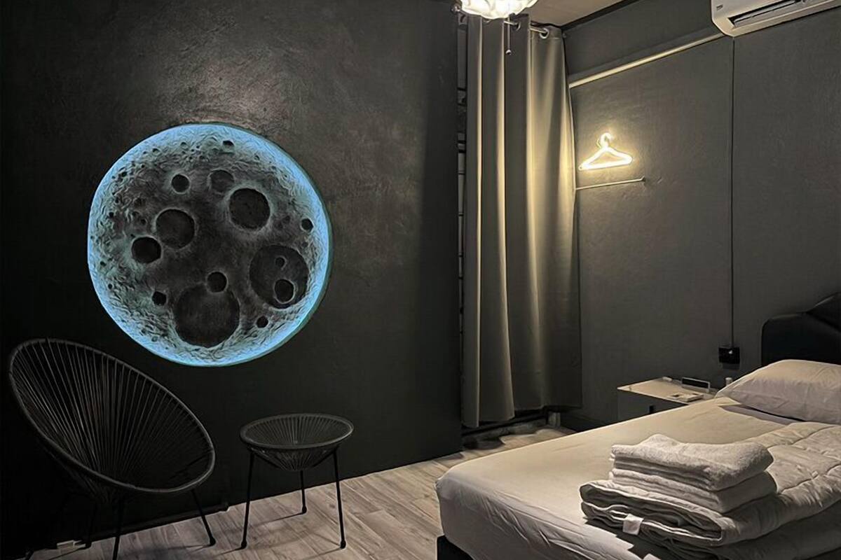 A bedroom with a large wall mural of the moon, a bed with neatly folded towels, a chair, and a hanging light fixture.