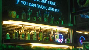 A bar area with a neon sign stating "WHY DRINK AND DRIVE IF YOU CAN SMOKE AND FLY" and various bottles of alcohol on shelves. - Doctor Alien Hostel (Bangkok)