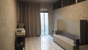 A room with a sofa, a small table, a trash bin, and a window with curtains. - Doctor Alien Hostel (Bangkok)