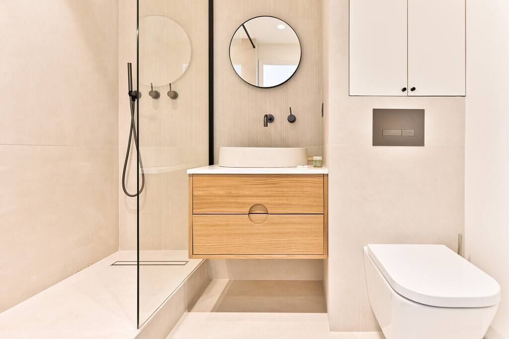Un baño moderno con cabina de ducha de vidrio, tocador de madera, espejo redondo y inodoro blanco.