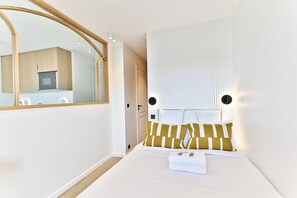 Quarto de hotel com uma cama, mesa de cabeceira, espelho, janela e vista para uma cozinha.