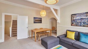 A living room with a sofa, ottoman, and a dining table with chairs. There is a staircase leading to another room. - Painters Apartment - modern decor, 4 bedrooms, central location. 5 minutes walk to South Bay beach. Fully equipped kitchen. Longer stays welcome. (North Yorkshire)