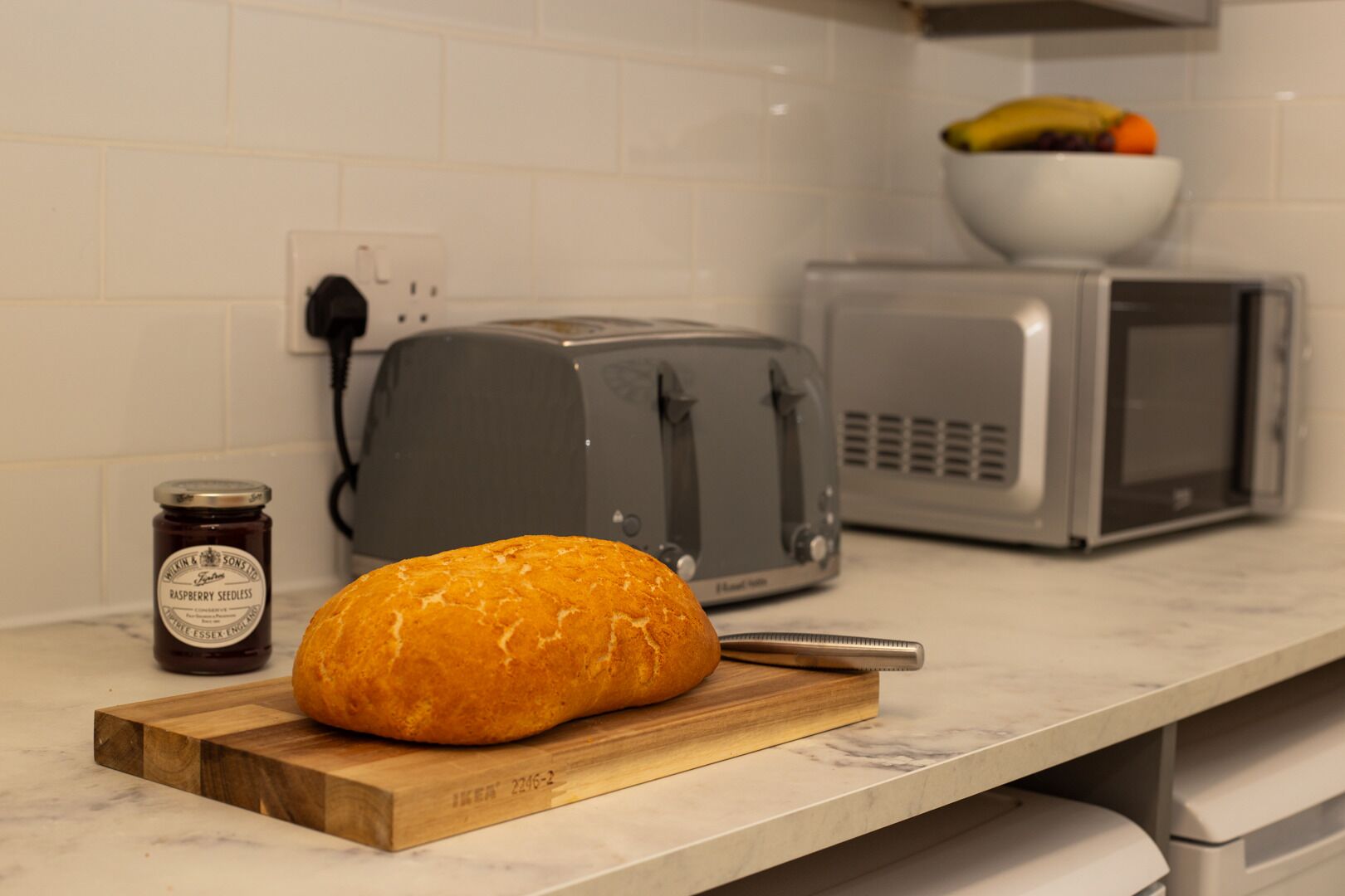 A kitchen with a loaf of bread on a wooden cutting board, a jar of raspberry seedless jam, a toaster, and a microwave.