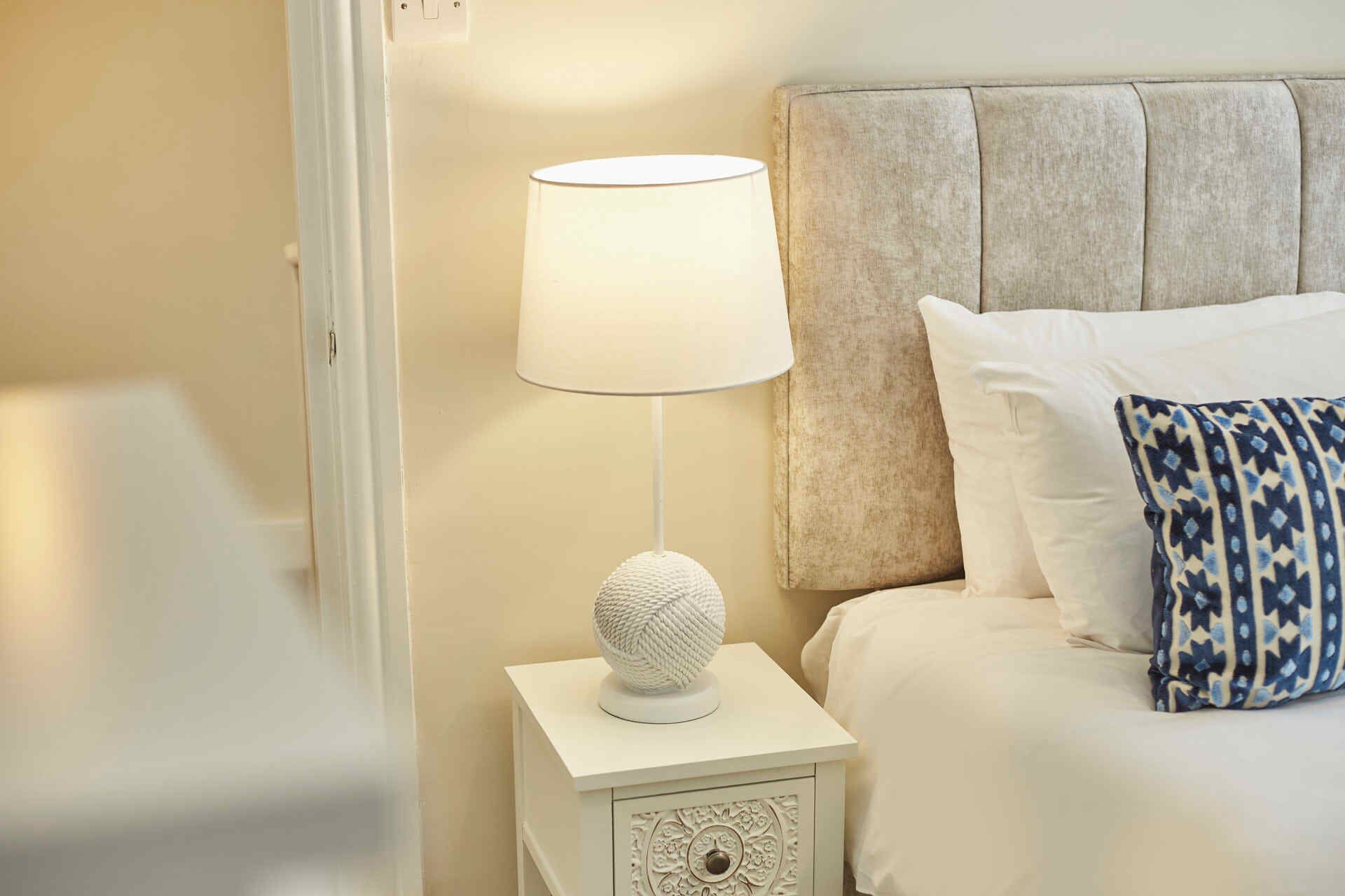 A neatly made bed with a patterned pillow, a white bedside lamp with a textured base, and a small bedside table with a decorative drawer.