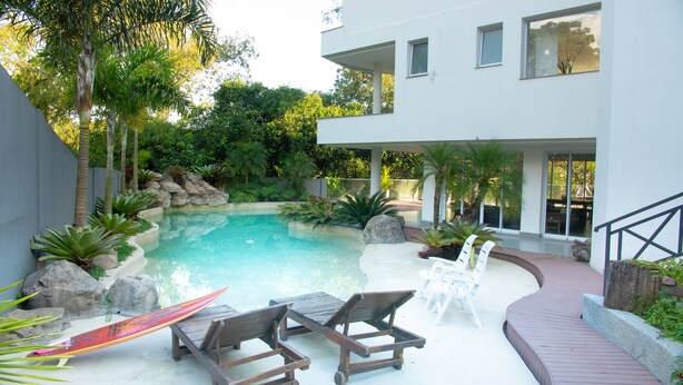 Ein modernes Haus mit Swimmingpool, umgeben von tropischen Pflanzen und einer Holzterrasse mit Liegestühlen.
