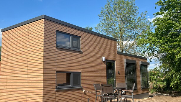Un bâtiment moderne à deux étages, doté d’une façade en bois, d’une terrasse avec mobilier d’extérieur et surplombant un ciel dégagé.