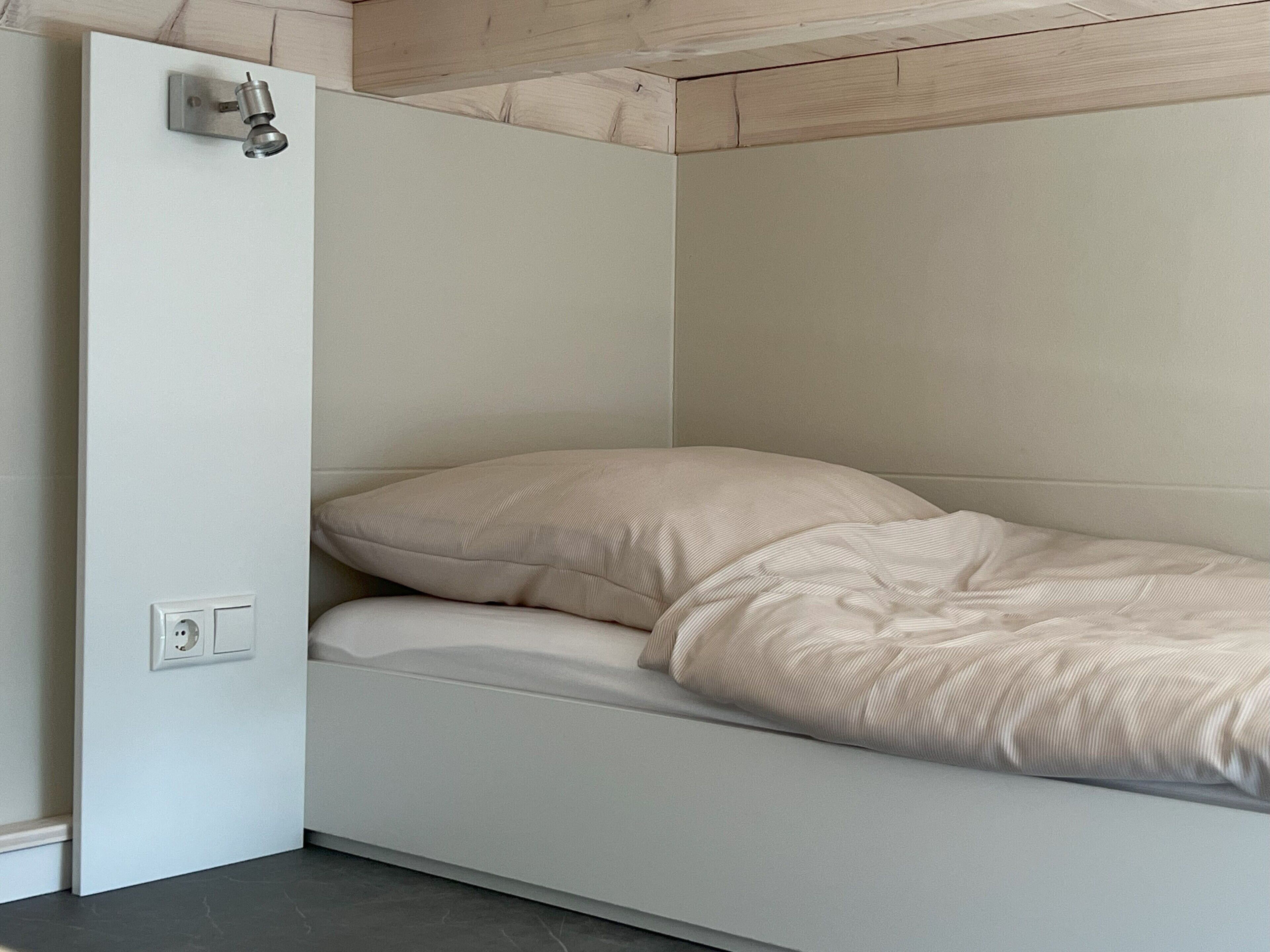 A compact bed with a headboard, a power outlet, and a light switch on the wall.