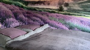 Une chambre avec une grande fresque murale représentant un champ de lavande au coucher du soleil, un lit avec une literie grise et un oreiller recouvert d’une housse violette.