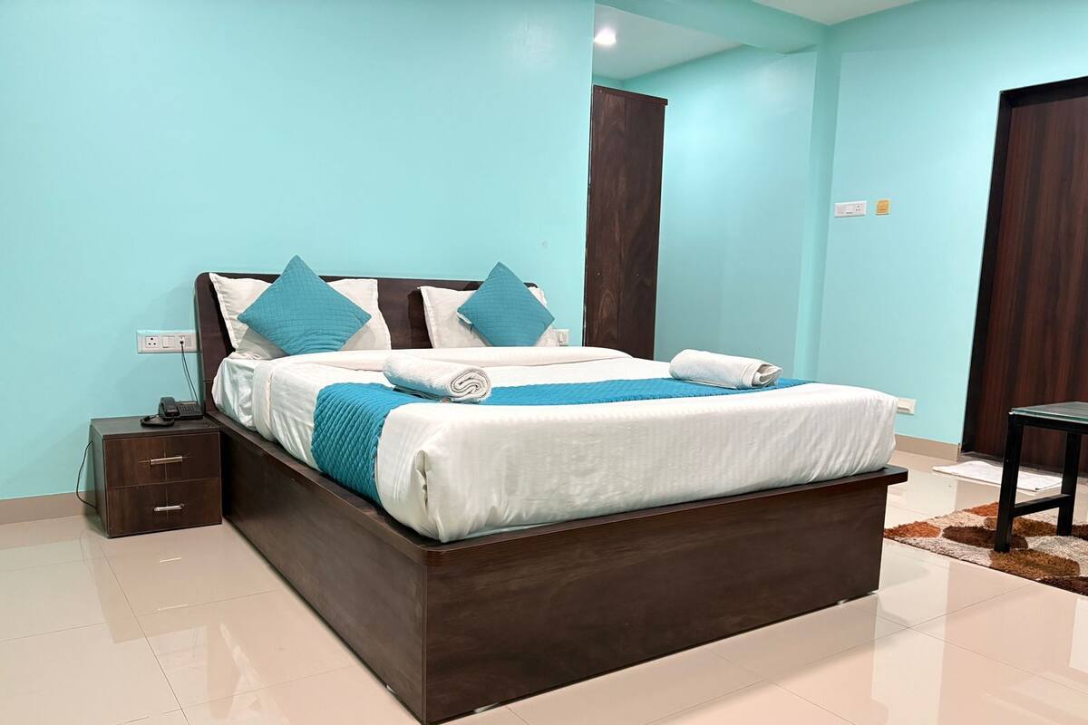 A neatly made bed with a dark wooden frame, a white mattress, and blue bedding. There is a small bedside table with a phone and charger, and a wall socket. The room has light blue walls and a tiled floor.