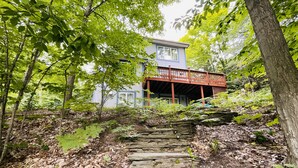 A stone staircase leading to a wooden deck with a house in the background surrounded by dense foliage. - Multi-week rental directly on Chandler Lake  deck, fire pit, tandem kayak. (Traverse City)