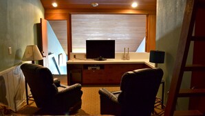 A room with a TV on a wooden cabinet, two leather chairs, a desk, and a ladder.