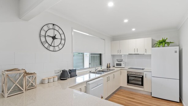 A modern kitchen with white cabinetry, a built-in oven, a microwave, a sink, and a large wall clock. - Renovated 3BR apartment with pool, WiFi, air-con & linen included, walking distance to beaches (Evans Head)
