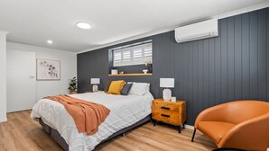 A bedroom with a bed, bedside tables, a chair, a window with blinds, an air conditioning unit, and a wall-mounted shelf. - Renovated 3BR apartment with pool, WiFi, air-con & linen included, walking distance to beaches (Evans Head)