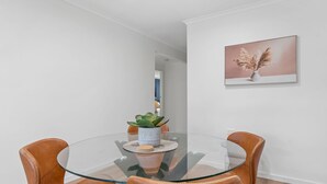 A modern dining area with a glass table, leather chairs, a potted plant, and a framed artwork on the wall. - Renovated 3BR apartment with pool, WiFi, air-con & linen included, walking distance to beaches (Evans Head)