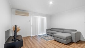 A modern living room with a grey sofa, a flat-screen TV, a wooden floor, and a large window with white curtains. - Renovated 3BR apartment with pool, WiFi, air-con & linen included, walking distance to beaches (Evans Head)