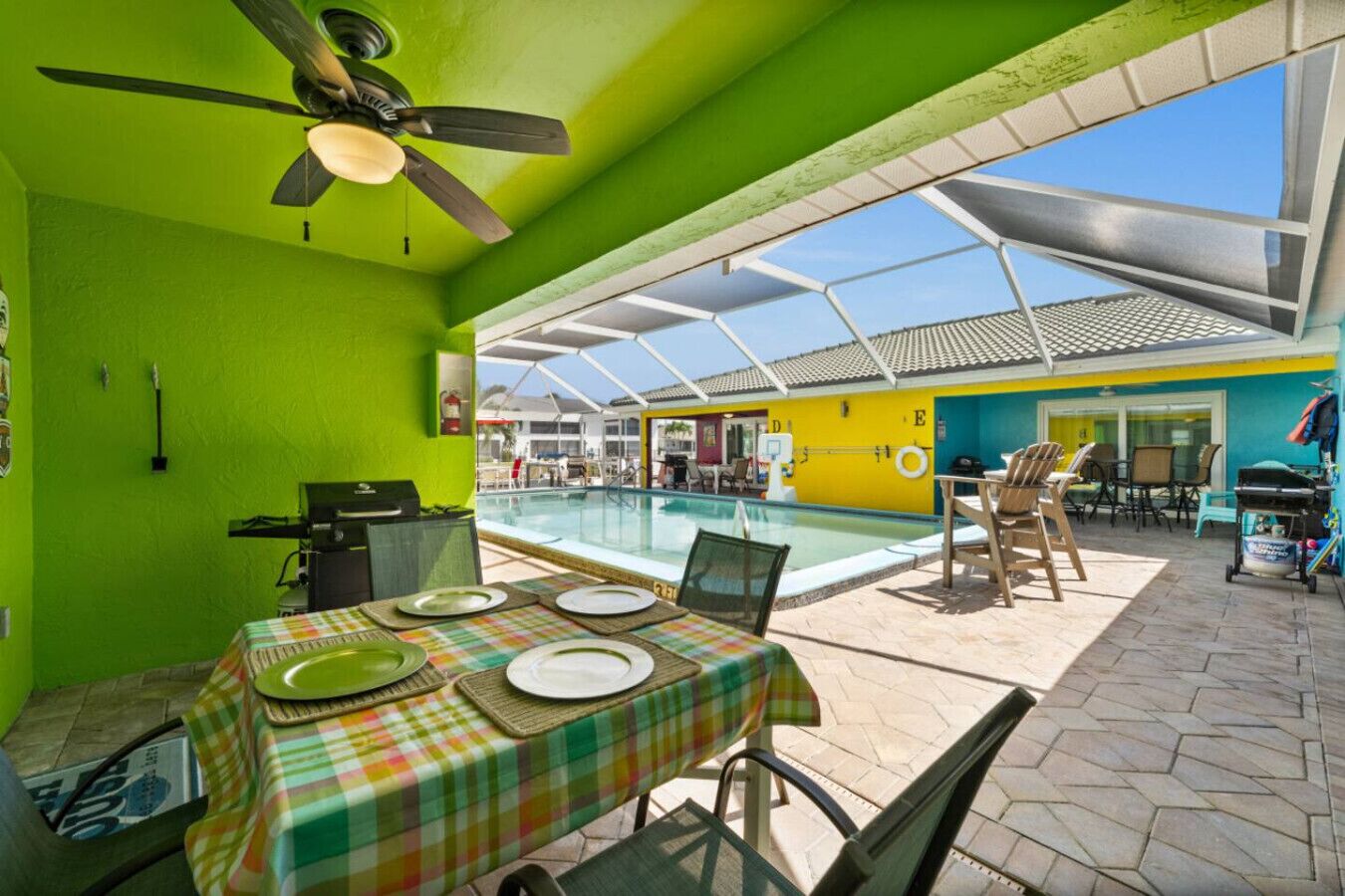 Zona de piscina con una mesa lista para comer, un ventilador de techo y vistas a la piscina y edificios cercanos.