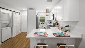 A modern kitchen with white cabinetry, a central island with bar stools, and a hanging pendant light. - Gulf Access Condo Heated Pool & Dock (Cape Coral)