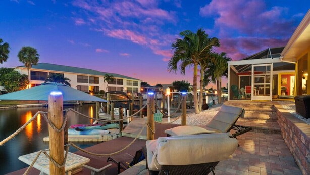 A waterfront patio with lounge chairs, a pool, and a building with a glass facade. - Gulf Access Condo Heated Pool & Dock (Cape Coral)