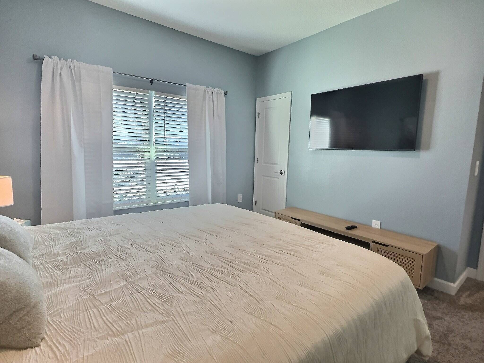 A bedroom with a bed, a TV mounted on the wall, a door, and a window with blinds.