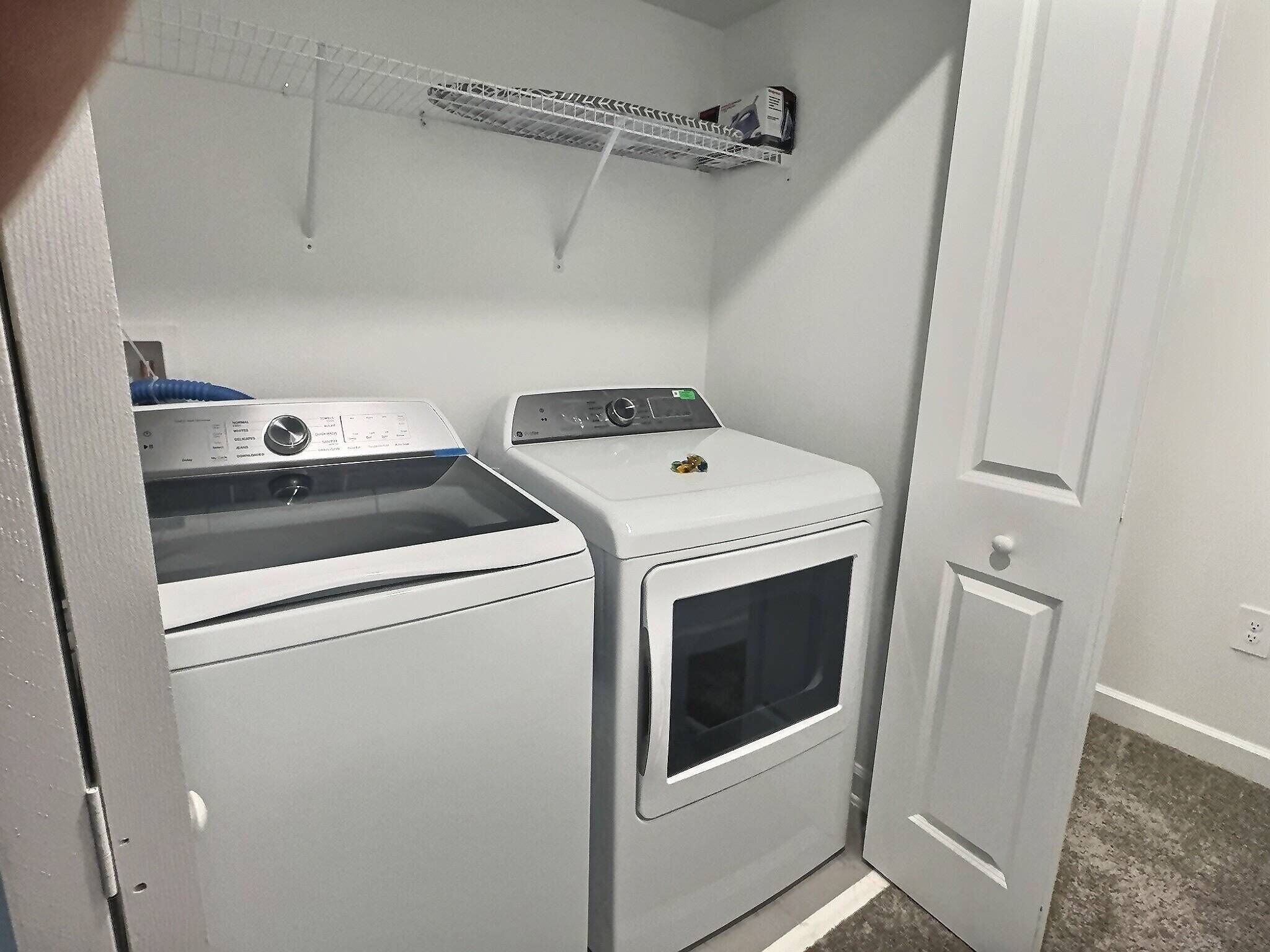 A compact laundry room with a front-loading washer and dryer, a small shelf above the dryer, and a door to the right.