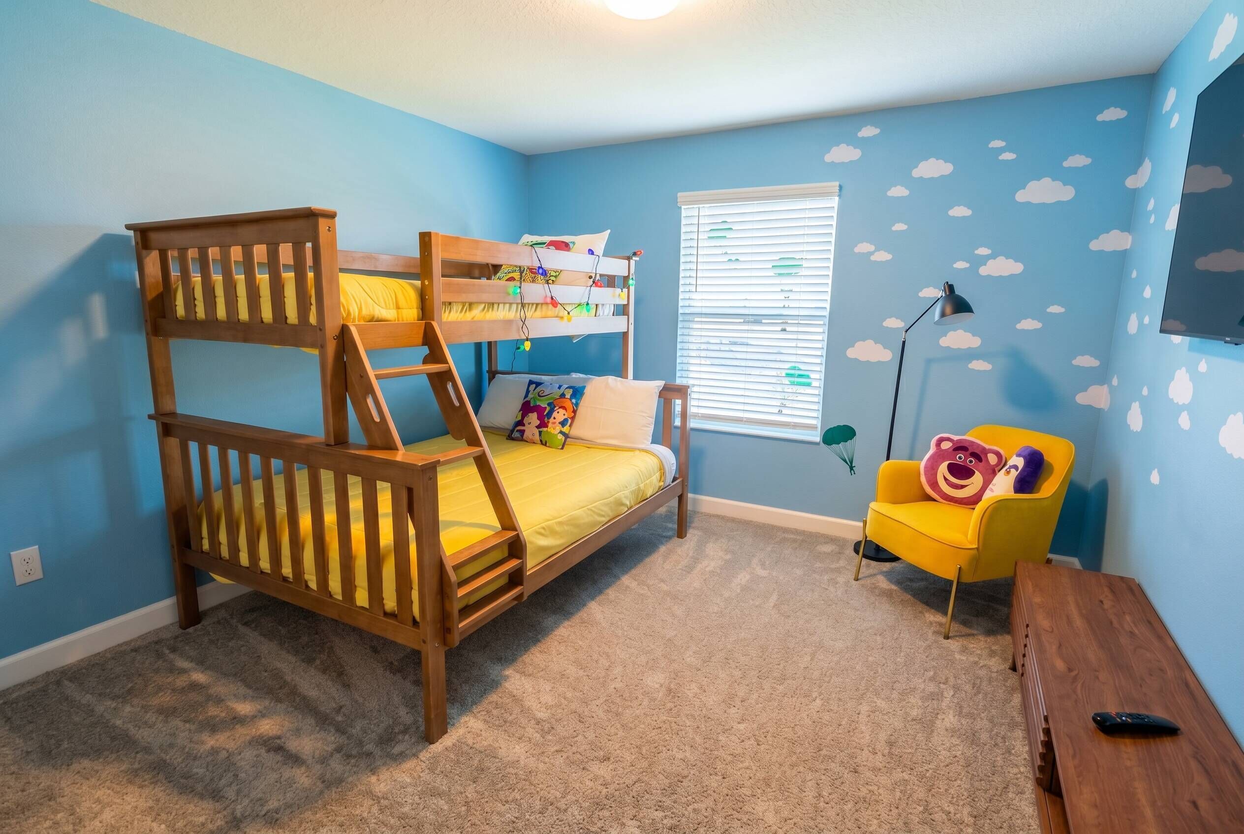 A bunk bed with a ladder, a yellow chair, a small table, a window with blinds, and a wall with cloud decals.