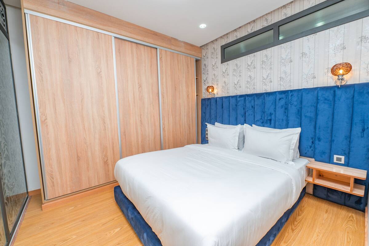 Een slaapkamer met een groot bed, een houten garderobe, een blauw hoofdeinde en twee wandlampen.