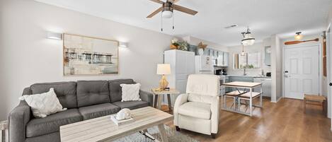 A living room with a grey sofa, a white armchair, a wooden coffee table, and a dining area with a small table and chairs.
