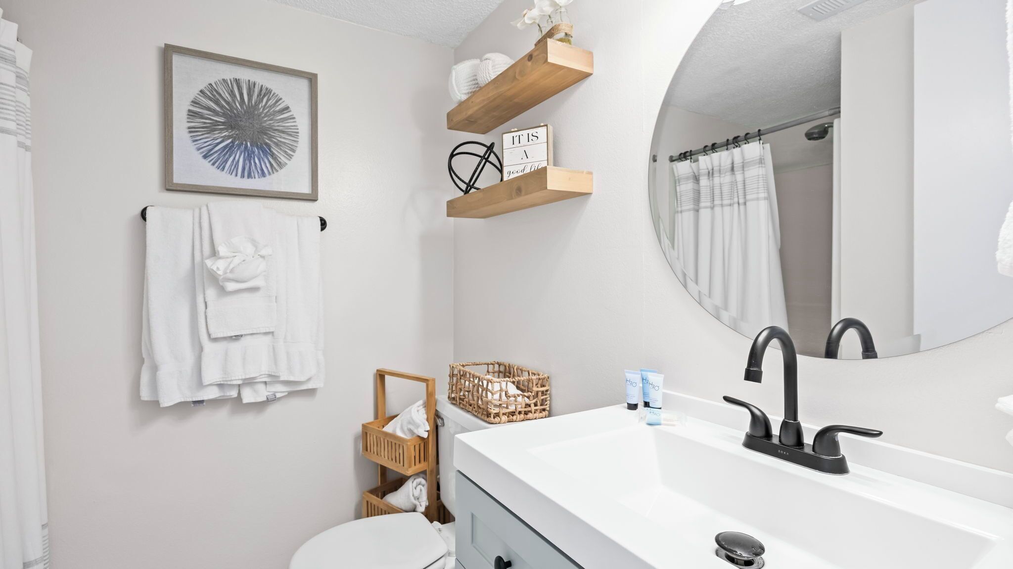 Un baño con un lavamanos blanco, grifo negro, una obra de arte abstracta enmarco, un toallero y un estante de madera con artículos de aseo.