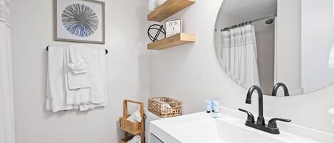 Un baño con un lavamanos blanco, grifo negro, una obra de arte abstracta enmarco, un toallero y un estante de madera con artículos de aseo.