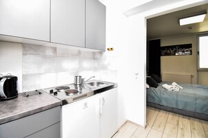 Una cocina compacta con diseño moderno, que incluye una estufa integrada, un hervidor y un lavamanos. Junto a la cocina se encuentra un dormitorio con una cama, almohadas y una ventana.