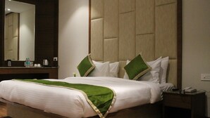 A hotel room with a bed, a bedside table, a nightstand, a mirror, and a wall-mounted panel. - Hotel Vedic Inn (Kiraoli)