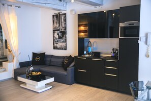 Cozinha moderna com armários pretos e eletrodomésticos de aço inoxidável, um sofá cinza com almofadas decorativas e uma mesa de centro branca com uma tigela e velas.