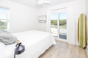 Et soveværelse med seng, ventilator, et vindue med persienner, en balkon med udsigt og et surfbræt.