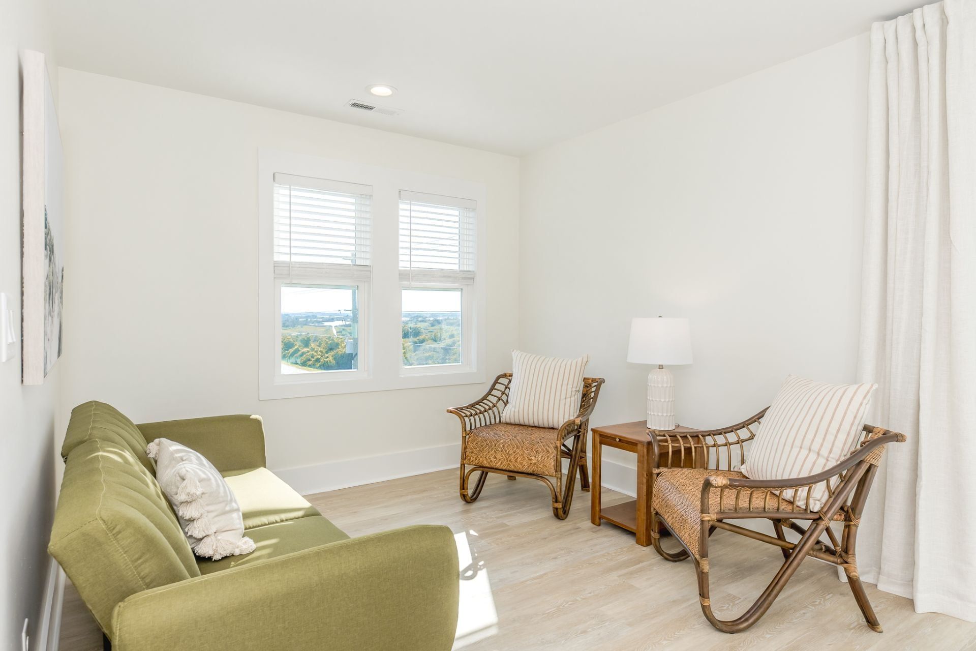 Une pièce avec un canapé vert, deux fauteuils en osier, une petite table avec une lampe et une fenêtre avec des stores.