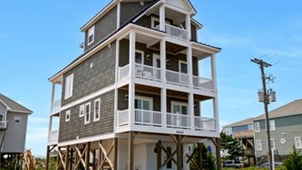 A multi-story building with a balcony on the ground floor, wooden support beams, and a clear sky. - In This Moment  - 5 BR New Oceanside Home; Elevator; Dog Friendly! (North Topsail Beach)
