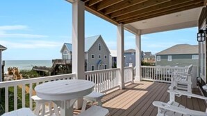 A covered outdoor deck with white furniture, overlooking a beach and residential buildings. - In This Moment  - 5 BR New Oceanside Home; Elevator; Dog Friendly! (North Topsail Beach)