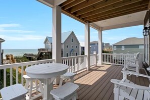 En overdækket terrasse med hvidt møbel, med udsigt over en strand og boliger.