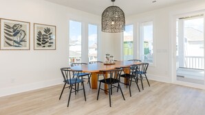 A spacious dining area with a wooden table and black chairs, large windows, and a hanging light fixture. - In This Moment  - 5 BR New Oceanside Home; Elevator; Dog Friendly! (North Topsail Beach)