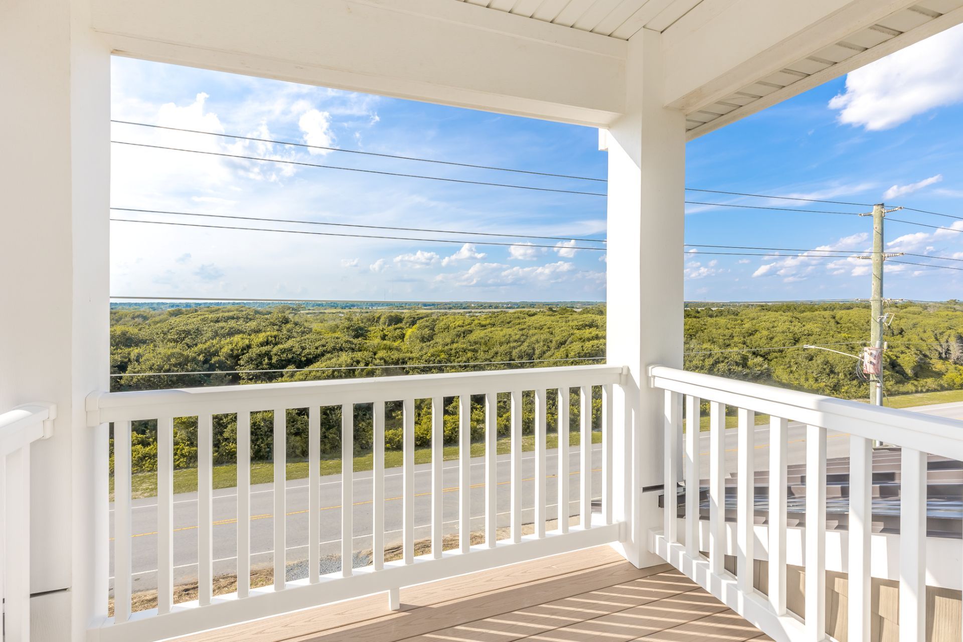 A covered balcony with a white railing, offering a view of a road and dense greenery.