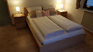 A neatly made bed with a white blanket and pillows, wooden headboard, and two bedside lamps on wooden nightstands. - Ferienwohnung Bohnert - Arias (Bad Peterstal-Griesbach)