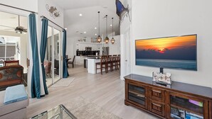A modern living room with a large TV, wooden furniture, and a view of the sunset. - Seagrape Retreat - Walking distance to the beach! (Marco Island)