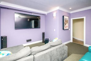 A living room with a large flat-screen TV mounted on the wall, a grey sectional sofa, and a teal chair. - Island Life - Amazing Oceanfront 9 BR Home with Pool, Theatre Room & More! (North Topsail Beach)