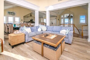 A spacious living room with a large sectional sofa, a coffee table with a chess set, wicker ottoman, and a view of the kitchen and dining area through an open archway. - Island Life - Amazing Oceanfront 9 BR Home with Pool, Theatre Room & More! (North Topsail Beach)