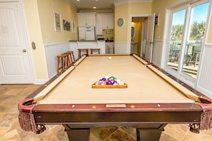 A pool table with balls in the center, surrounded by a room with a kitchen and a view through large windows. - Island Life - Amazing Oceanfront 9 BR Home with Pool, Theatre Room & More! (North Topsail Beach)