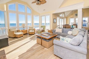 A spacious living room with large windows, a ceiling fan, and a chessboard on a coffee table. - Island Life - Amazing Oceanfront 9 BR Home with Pool, Theatre Room & More! (North Topsail Beach)