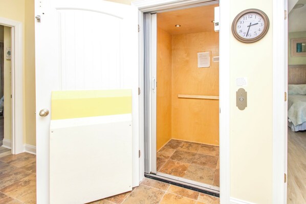 An elevator with a yellow safety line, a clock on the wall, and a door leading to a room with a bed.