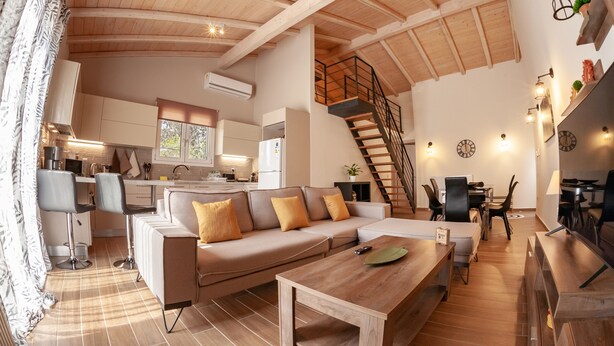 A spacious living area with a high ceiling, a staircase, a sofa with cushions, a wooden table, and a kitchen in the background.