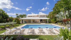 Una casa grande con piscina, rodeada de plantas tropicales y un cielo azul despejado.