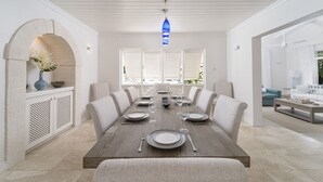 Comedor con una larga mesa dispuesta para seis personas, paredes blancas y un ventanal con contraventanas.