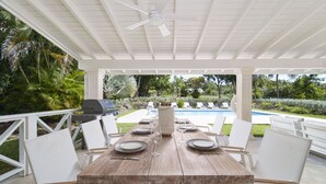 Un espacio para comer al aire libre con una mesa de madera y sillas blancas, un ventilador de techo y vistas a una piscina y zonas verdes.